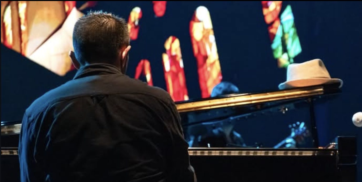 César Ibacache al piano de cola en concierto, de espaldas frente a la proyección del escenario